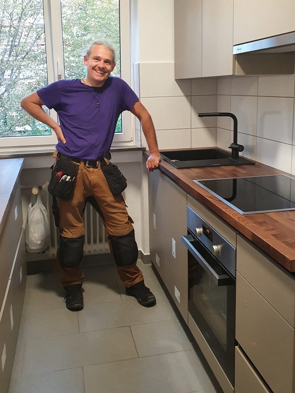 Tilman Sandkühler in Arbeitskleidung mit Werkzeugtasche, lächelnd in einer fertig montierten Küche mit Holzarbeitsplatte.