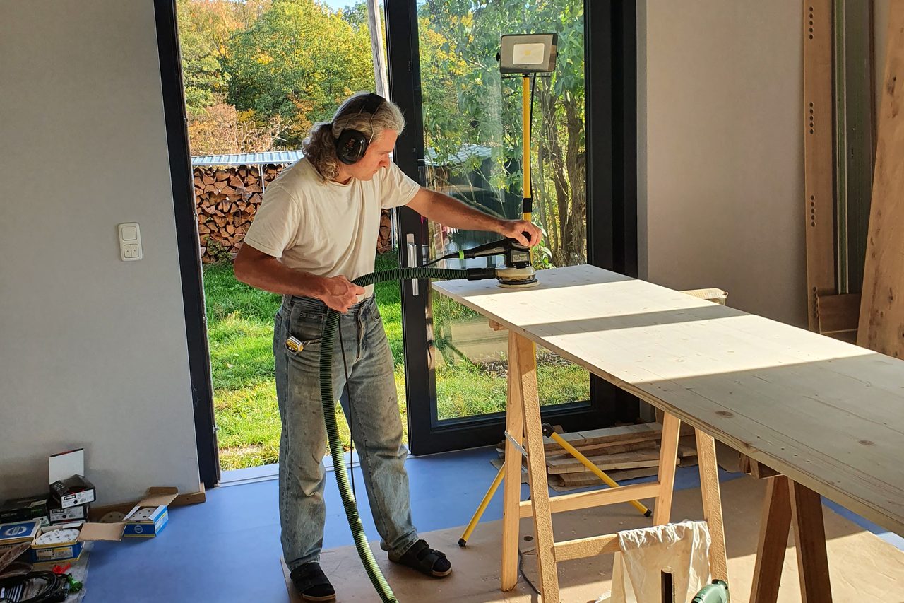 Tilman Sandkühler schleift mit einer Schleifmaschine eine Holzplatte auf zwei Holzböcken, dahinter eine offene Terrassentür mit Blick in den Garten.