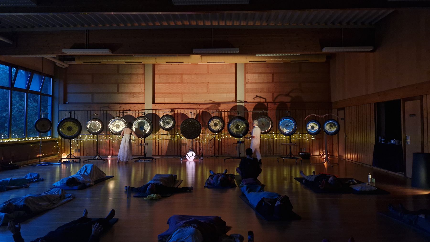 Klangveranstaltung in einer Halle: Eine lange Reihe von Gongs, davor zwei stehende Personen, Teilnehmende liegen auf Matten.