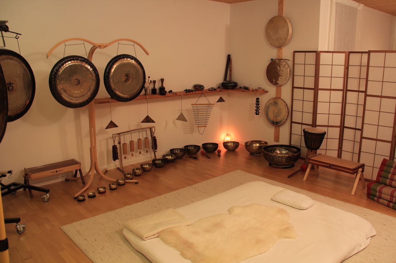 Der Klangraum: Gongs und Klangschalen an der Wand und auf dem Boden, davor ein vorbereiteter Liegeplatz mit Schaffell, warmes Licht.