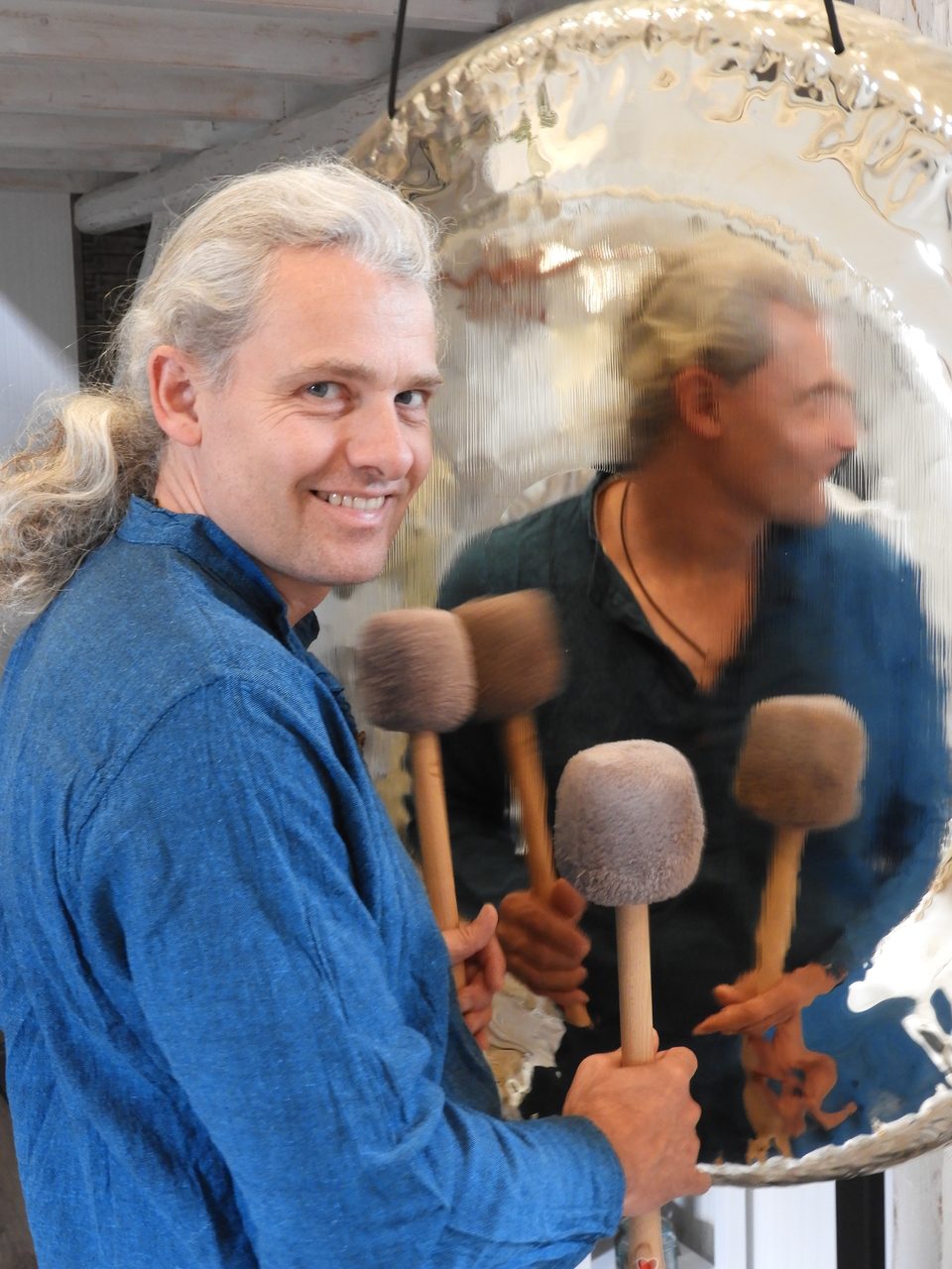 Tilman Sandkühler lächelt in die Kamera und hält mehrere Gongschlägel vor einem großen, spiegelnden Gong, in dem sich sein Spiegelbild zeigt.