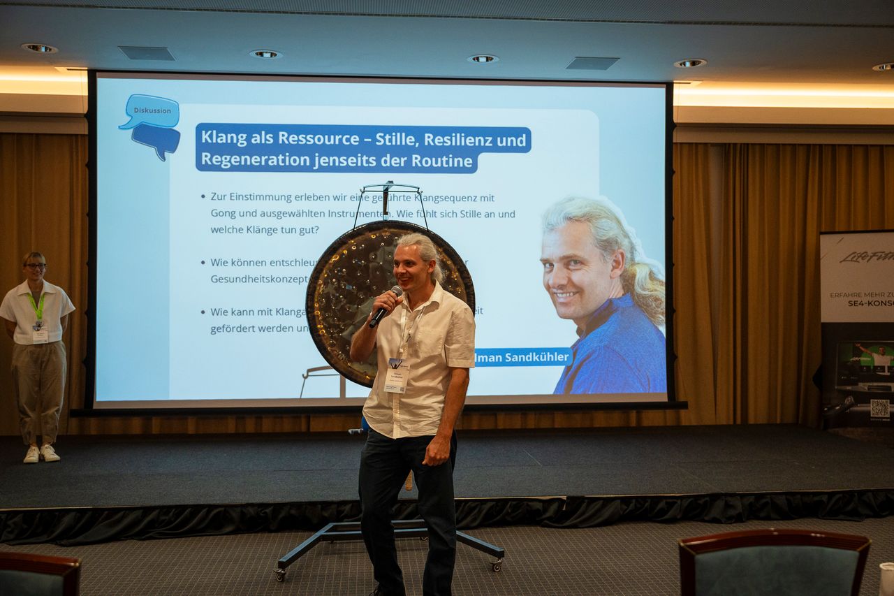 Tilman Sandkühler spricht mit Mikrofon vor einem Gong, hinter ihm die Präsentation „Klang als Ressource“ auf der Leinwand.